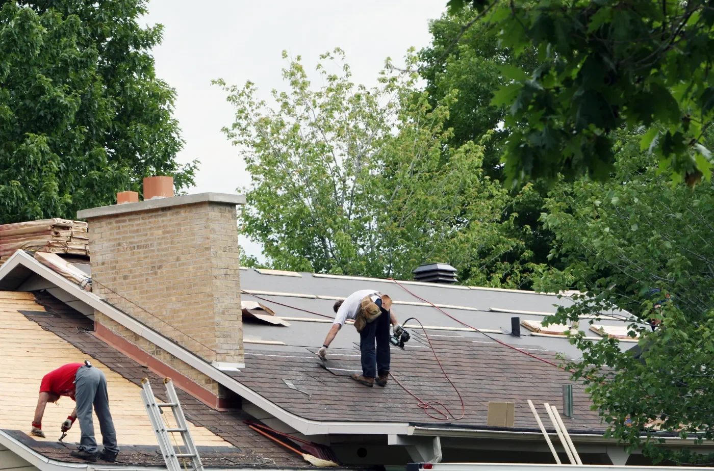 Two roofers are working. One with drill and shingles. Other with nail and hammer