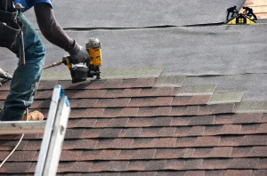Worker installing brown asphalt shingles on a roof using a nail gun.