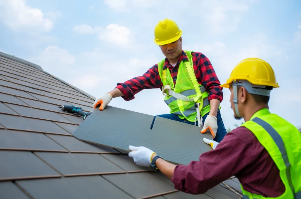 Two construction workers in safety gear placing a sheet of roofing material onto a sloped roof.