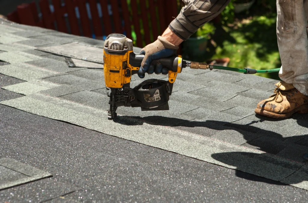 Worker using a nail gun to install asphalt shingles on a roof.