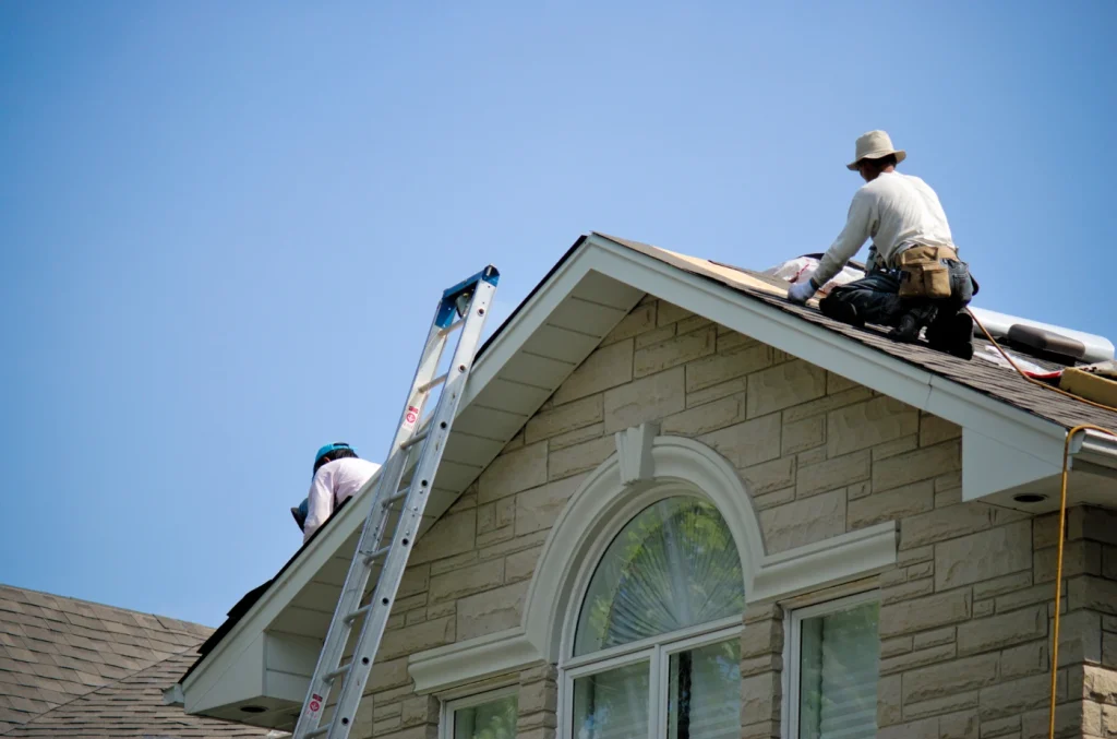 Workers installing roofing materials on a light-colored house under a clear blue sky, with a ladder positioned against the roof.