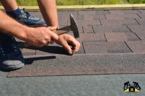 Close-up of a worker’s hands installing asphalt shingles on a roof using a hammer.