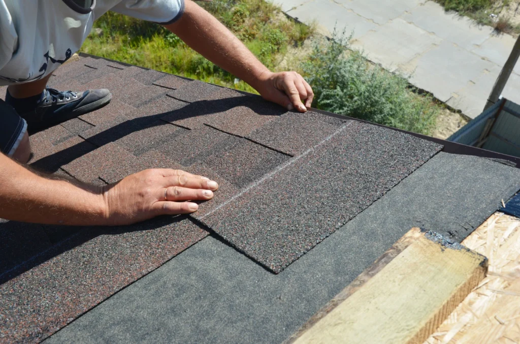 Worker positioning roofing materials on a roof surface, with asphalt shingles and underlayment visible.
