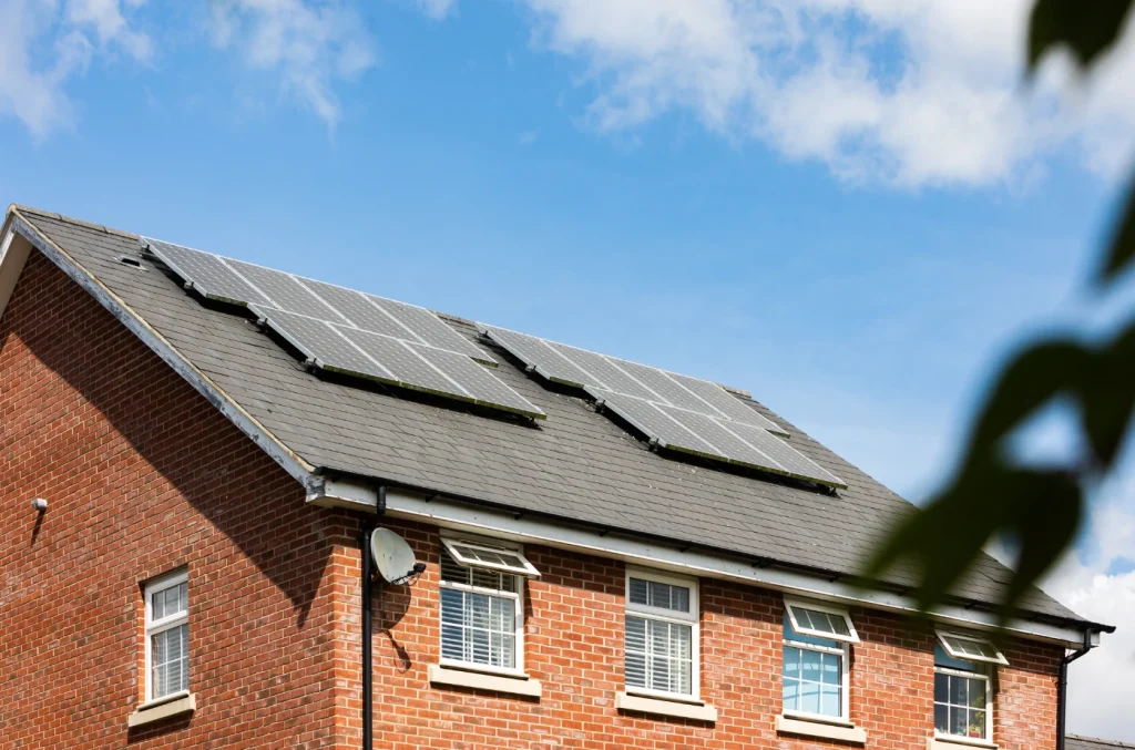 Close-up view of a residential house roof with dark shingles against a blue sky, showing the roofline and part of the brick exterior.